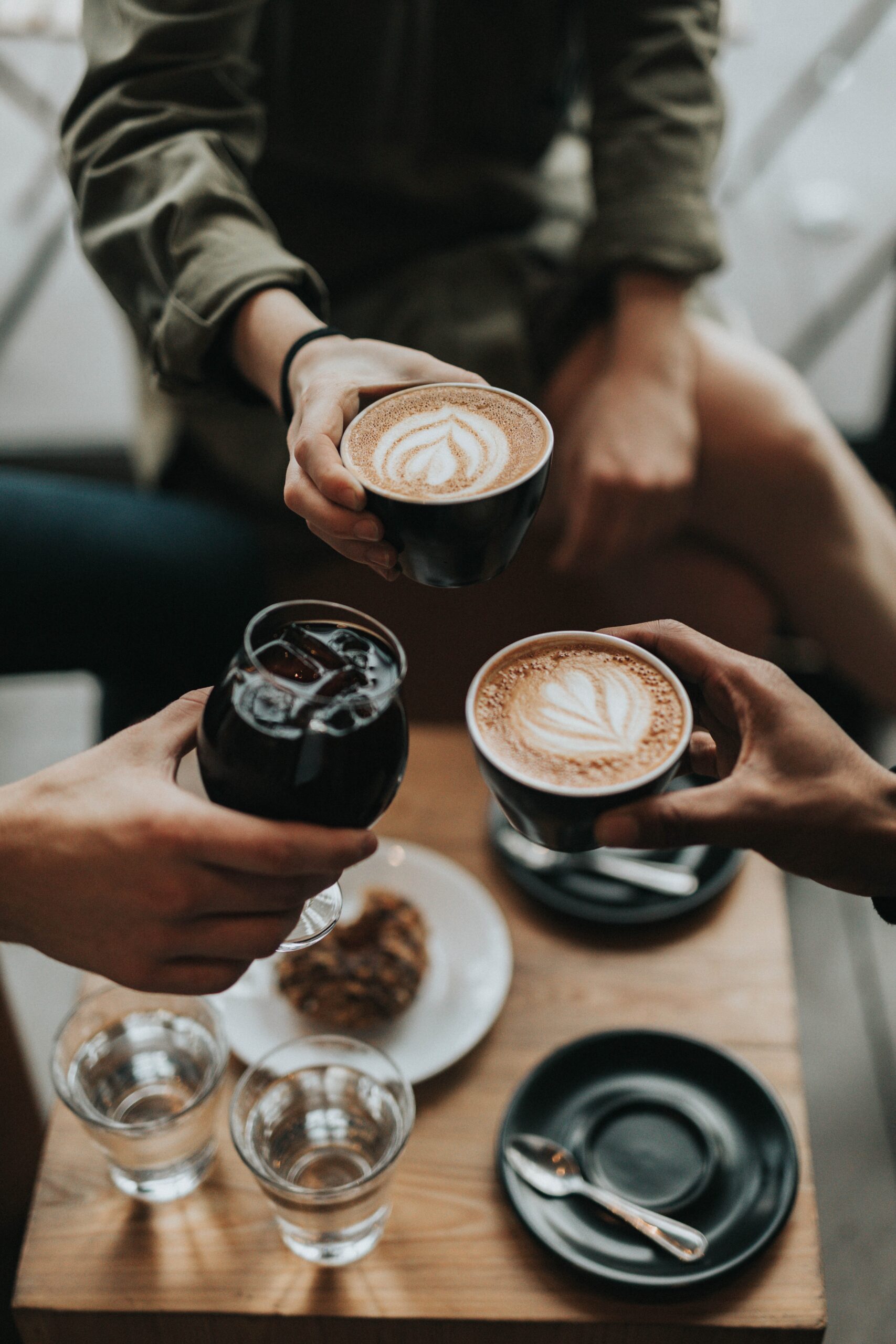 A birds eye view of three people lifting their drinks to cheers each other. Two have latte's and one has a coke.