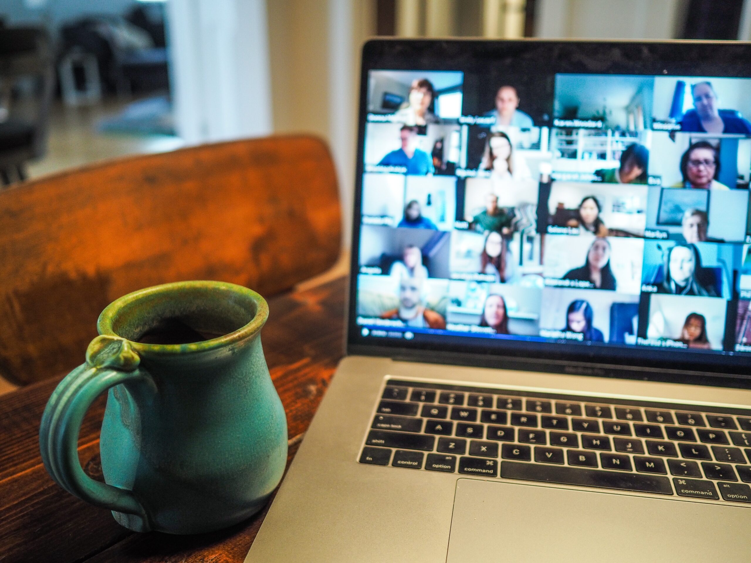 A large zoom meeting on a mac laptop. A green coffee mug sits next to the laptop.