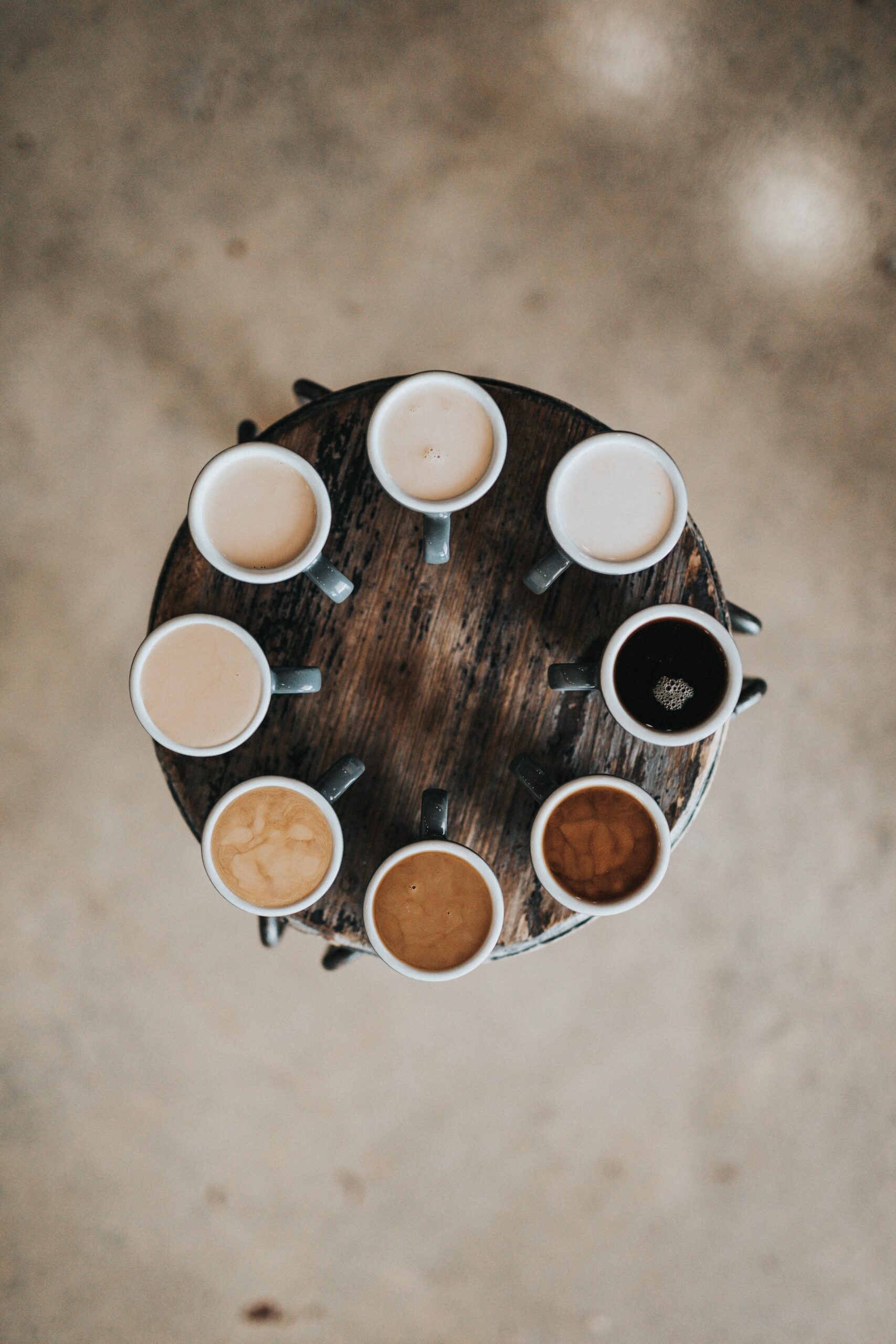 A circular wooden table with eight white mugs of tea around its edge. They're handles are pointed in and the tea gets darker within each mug until you get a tea that has no milk in it.
