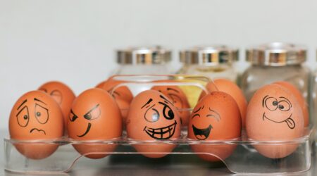 A group of 6 eggs in a clear container, each egg has a funny face drawn on it.