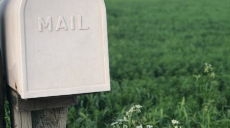 A cream mailbox on a weathered wooden post in the left foreground. The rest of the photo shows a green field with a line of trees and then the sky in the upper half of the background.